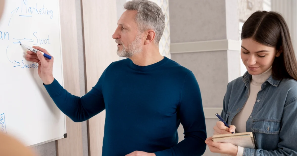 Man writing on whiteboard while woman takes notes.
